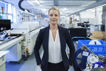 Confident smiling businesswoman standing with hands in pockets in illuminated factory - MOEF03413