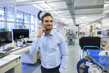 Smiling young male professional talking on smart phone while standing at illuminated factory - MOEF03418