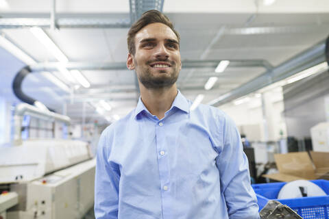 Lächelnder junger männlicher Ingenieur, der in einer beleuchteten Fabrik steht und wegschaut, lizenzfreies Stockfoto