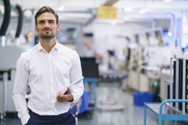 Confident young businessman holding digital tablet while standing at illuminated factory - MOEF03438