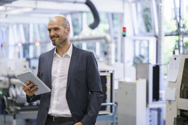 Lächelnder Geschäftsmann, der ein digitales Tablet hält und in eine helle Fabrik schaut - MOEF03440