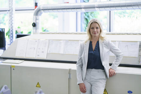 Confident blond businesswoman standing against machinery in illuminated factory stock photo