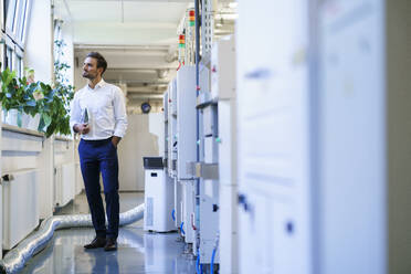 Thoughtful male technician holding digital tablet while looking away at illuminated factory - MOEF03451