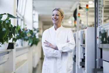 Confident mature female scientist with hands in lab coat pockets standing by machinery at laboratory - MOEF03457