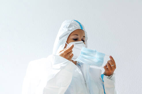 Krankenschwester in Arbeitsschutzkleidung mit Gesichtsmaske, die an einer weißen Wand steht - PGF00092