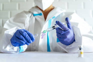 Krankenschwester mit Schutzanzug füllt eine Injektion ab, während sie am Tisch im Krankenhaus sitzt - PGF00095