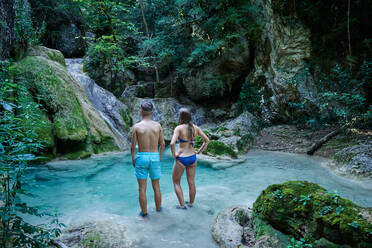 Rückansicht eines Paares von Reisenden in Badekleidung, die im klaren Wasser eines Sees in den Bergen stehen und die herrliche Landschaft mit Felsen genießen - ADSF15680