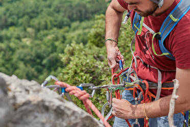 High angle of crop male alpinist in safety harness hanging on rope on rock and adjusting carabiners - ADSF15689