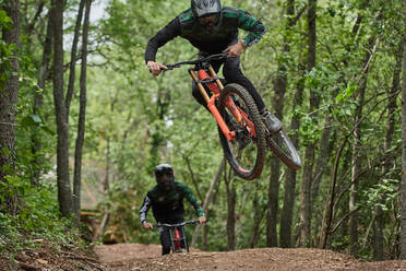 Male cyclists in protective helmets and costumes performing dangerous stunts on bikes for downhill on trail in forest - ADSF15693