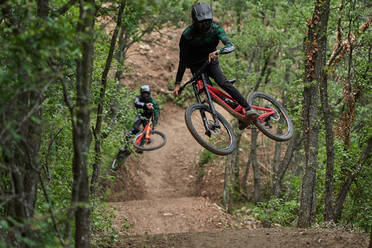 Männliche Radfahrer in Schutzhelmen und Kostümen, die gefährliche Stunts auf Fahrrädern beim Downhill auf einem Pfad im Wald vollführen - ADSF15695