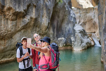 Gruppe von begeisterten Freunden mit Rucksäcken, die zusammen ein Selfie mit ihrem Smartphone machen, während sie in einer Schlucht in der Nähe eines Sees stehen, während der Sommerferien - ADSF15700