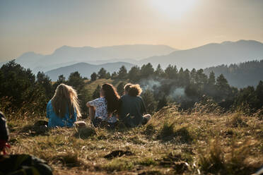 Back view of group of unrecognizable travelers resting on grass and enjoying sunset in mountains during summer adventure - ADSF15701