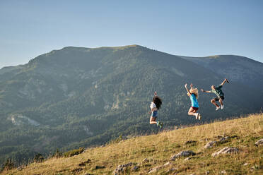 Company of delighted friends jumping on hill in mountains while enjoying freedom during summer vacation - ADSF15703