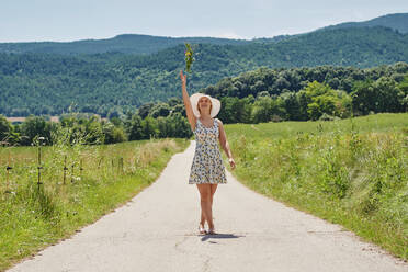 Zufriedene Frau in Kleid und Sonnenhut, die mit einem Blumenstrauß auf der Straße vor dem Hintergrund der Berge spazieren geht und nach oben schaut - ADSF15705