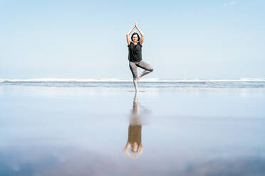 Junge Frau in Sportkleidung steht in Baumstellung mit erhobenen Armen, während sie Yoga am Sandstrand gegen das wogende Meer mit geschlossenen Augen praktiziert - ADSF15769