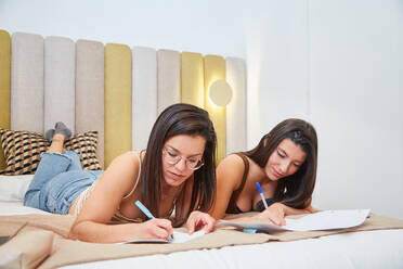 Clevere Studentinnen liegen auf dem Bett und schreiben Notizen auf Papier, während sie gemeinsam Hausaufgaben machen - ADSF15809
