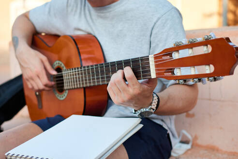 Kreativer männlicher Musiker, der auf einer Steinbank auf der Straße sitzt und Noten liest, während er sich auf das Gitarrenspiel vorbereitet - ADSF15811