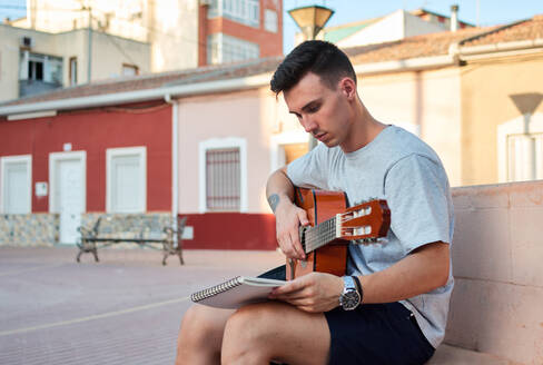 Kreativer männlicher Musiker, der auf einer Steinbank auf der Straße sitzt und Noten liest, während er sich auf das Gitarrenspiel vorbereitet - ADSF15812