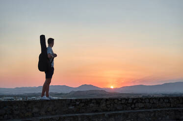 Seitenansicht eines nicht erkennbaren männlichen Gitarristen, der mit einem Musikinstrument im Koffer auf einem Steinzaun steht und den herrlichen Sonnenuntergang in den Bergen bewundert - ADSF15813