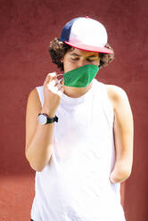 Teenage boy wearing face mask while standing against wall on sunny day - JCMF01524