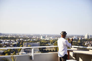 Älterer Mann, der ein Foto macht, während er sein Smartphone benutzt und auf der Terrasse eines Gebäudes steht - FMKF06326