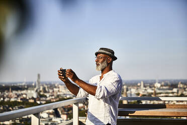 Älterer Mann, der ein Selfie macht, während er ein Smartphone benutzt, das auf einer Gebäudeterrasse steht - FMKF06327