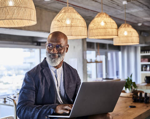 Smiling man looking away while working on laptop sitting at home stock photo