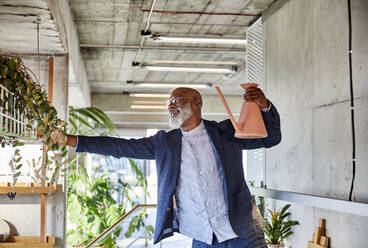 Älterer Mann hält eine Gießkanne und betrachtet eine Pflanze, während er zu Hause steht - FMKF06357