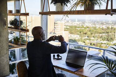 Älterer Mann, der ein Foto mit seinem Smartphone macht und durch das Fenster schaut, während er zu Hause sitzt - FMKF06373