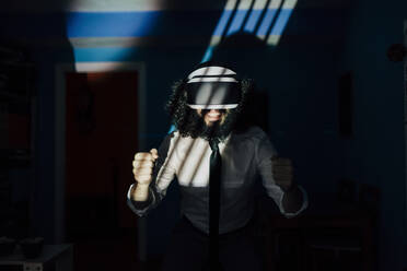 Playful man wearing virtual reality glasses while standing in dark at home during pandemic - MRRF00488