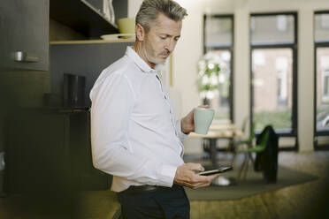 Geschäftsmann hält Tasse, während er ein Mobiltelefon im Büro benutzt - JOSEF01957