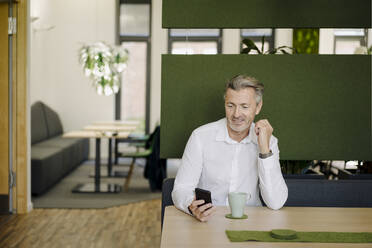 Smiling businessman using mobile phone while sitting on table at cafeteria - JOSEF01961