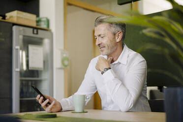Mann lächelt, während er am Tisch in einer Cafeteria sitzt und ein Mobiltelefon benutzt - JOSEF01963