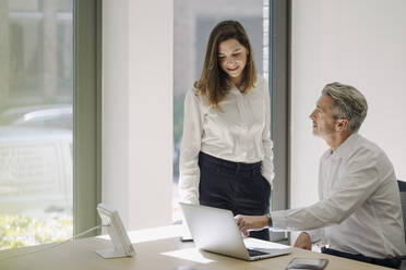 Businessman pointing at laptop while sitting on chair by woman at office - JOSEF01965