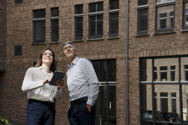 Businessman and woman looking up while using digital tablet against building - JOSEF01973