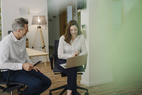 Smiling woman using laptop while sitting on chair by businessman in office stock photo