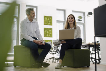 Lächelnde Partner, die einen Laptop benutzen, während sie im Büro sitzen - JOSEF01989