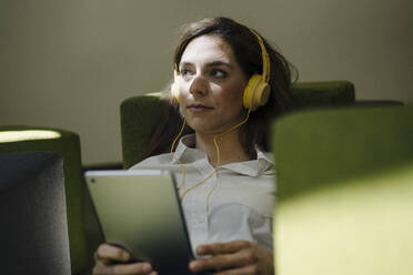 Frau mit Kopfhörern, die wegschaut, während sie ein digitales Tablet im Büro benutzt - JOSEF02000