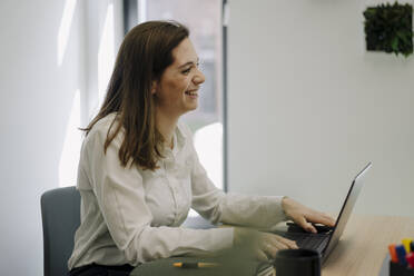 Lächelnde Geschäftsfrau, die einen Laptop benutzt, während sie im Büro sitzt - JOSEF02004