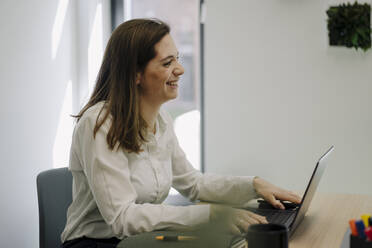 Smiling businesswoman using laptop while sitting in office - JOSEF02004