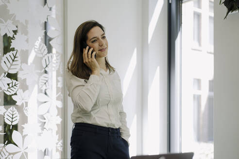 Geschäftsfrau mit Hand in der Tasche, die im Büro stehend mit ihrem Mobiltelefon spricht - JOSEF02006