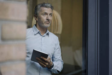 Geschäftsmann schaut durch ein Fenster, während er im Büro ein digitales Tablet benutzt - JOSEF02014