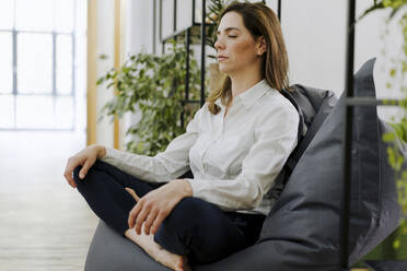 Woman practicing yoga while sitting on bean bag at office - JOSEF02039