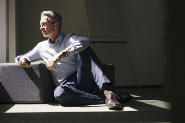 Businessman sitting by hassock on floor at office - JOSEF02063