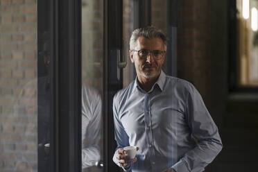 Businessman wearing eyeglasses holding coffee cup while leaning on window at office - JOSEF02075