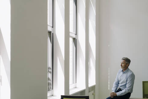 Geschäftsmann schaut durch das Fenster, während er auf dem Tisch im Büro sitzt - JOSEF02084