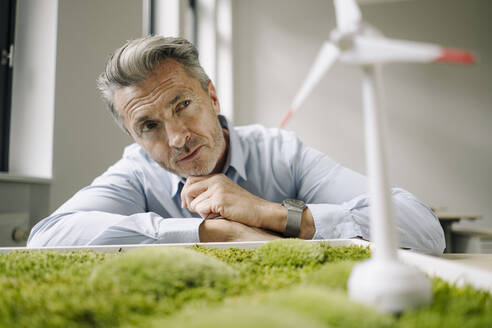 Businessman looking at wind turbine toy over moss frame while leaning on table at office - JOSEF02111