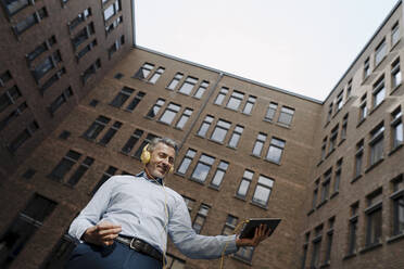 Happy man with headphones and digital tablet dancing while standing against building exterior - JOSEF02120