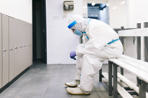 Thoughtful man wearing protective suit sitting on bench in hospital - PGF00112