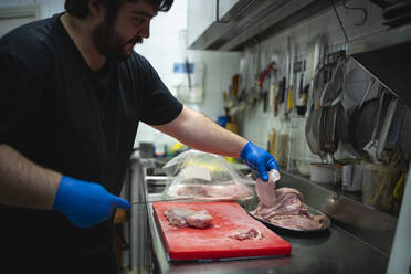 Koch schneidet Fleisch auf einem Schneidebrett, während er in einer Großküche steht - SNF00533